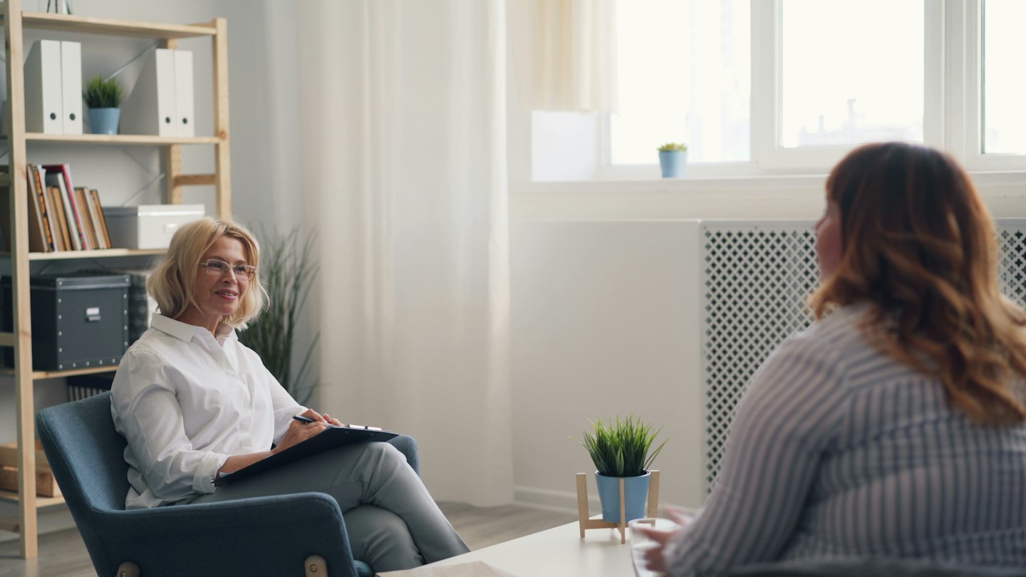 Woman in teal chair with clipboard listens to client.